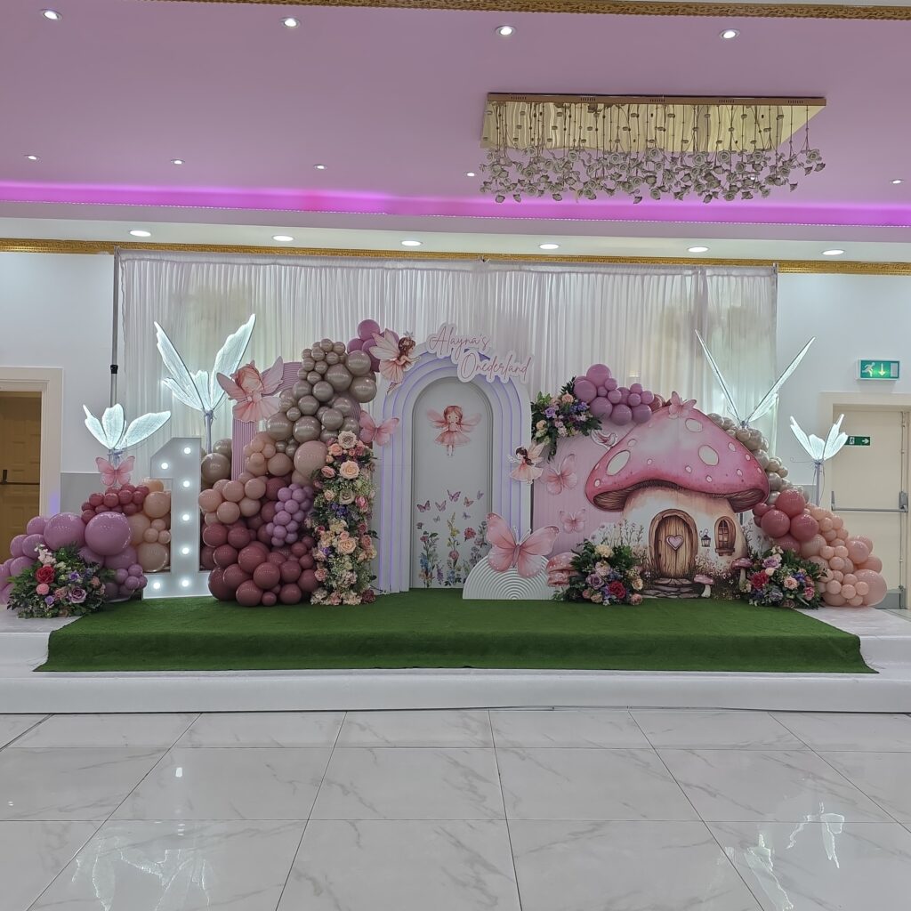 Wonderland-themed first birthday decor with pastel balloon garlands, a fairy-tale mushroom house, glowing fairy wings, and floral arrangements.