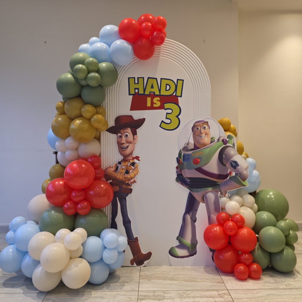 Toy Story-themed birthday backdrop with Woody and Buzz Lightyear cut-outs, colourful balloon arch, and "Hadi is 3" sign.