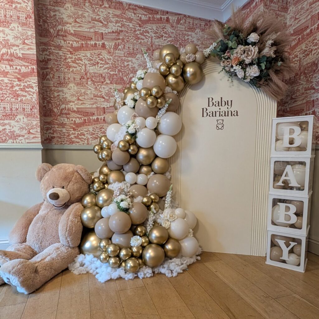 Neutral and gold baby shower decor with balloon garland, floral arrangement, teddy bear, and stacked “BABY” letter boxes.