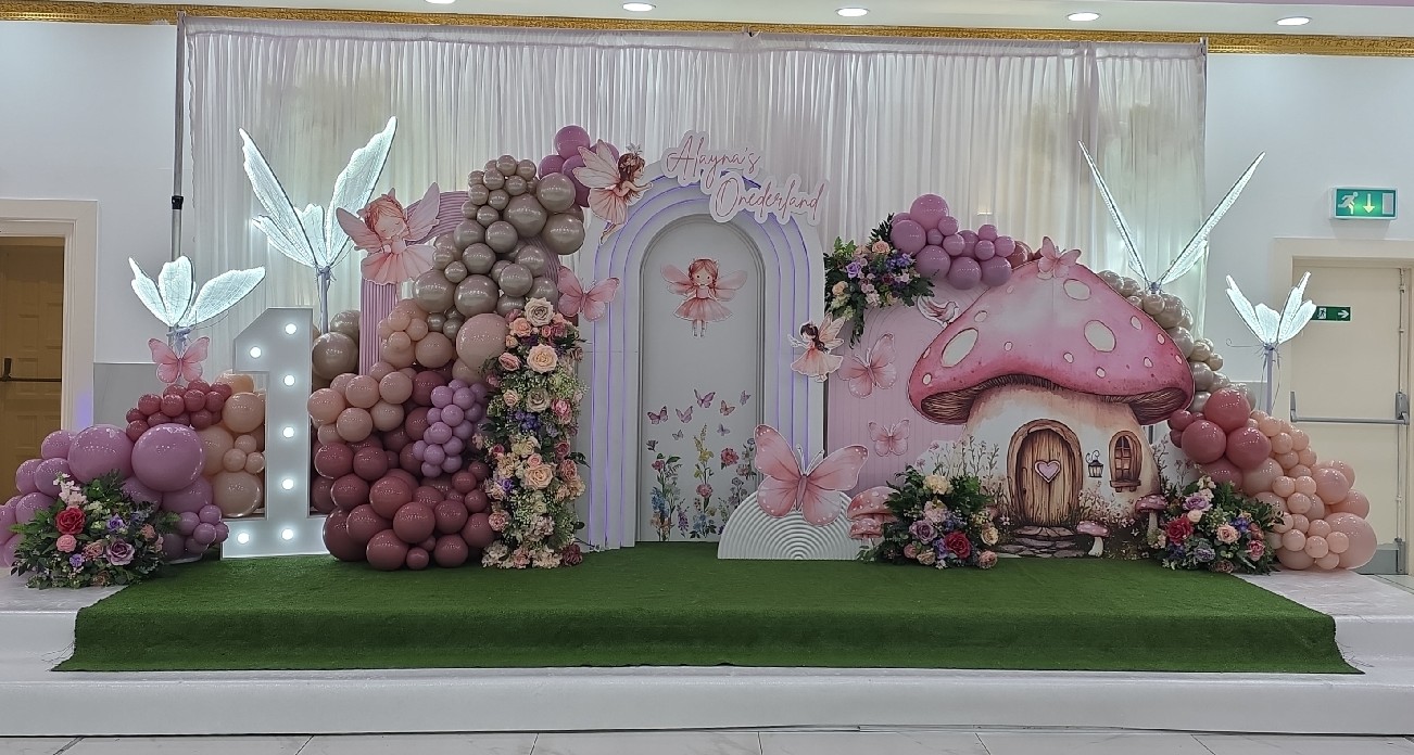 Whimsical fairy-themed first birthday setup featuring pastel balloon clusters, a light-up number one, floral arrangements, butterfly and fairy cutouts, and a toadstool house backdrop with “Aayna’s Wonderland” signage.