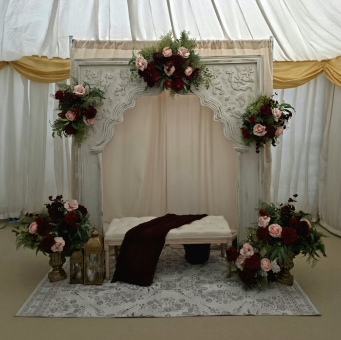 Luxury burgundy and blush Nikkah setup with carved arch, floral arrangements and upholstered bench in London
