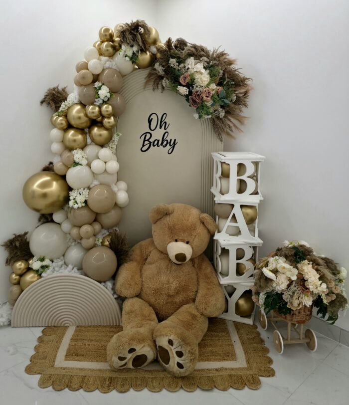 Baby shower display with beige and gold balloon garland, teddy bear, floral accents and Oh Baby backdrop.