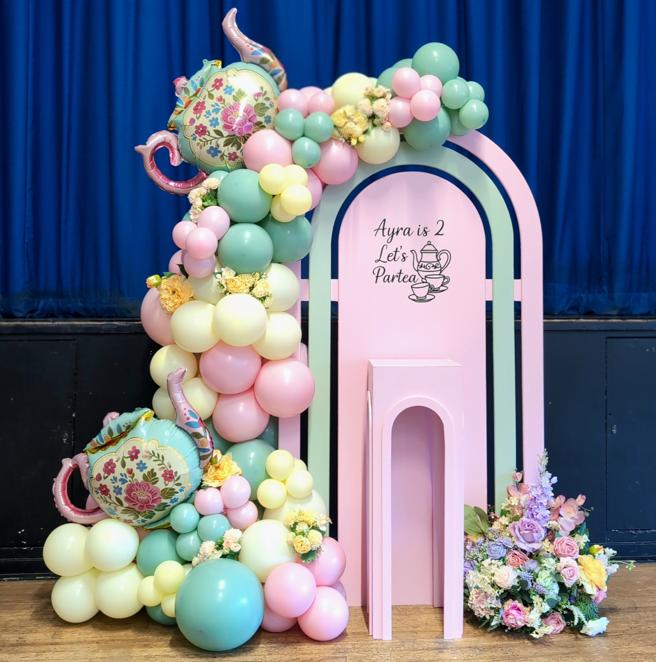 Tea party themed birthday decor with pastel balloons, arch backdrop and fresh flowers arranged beside a pink display plinth.