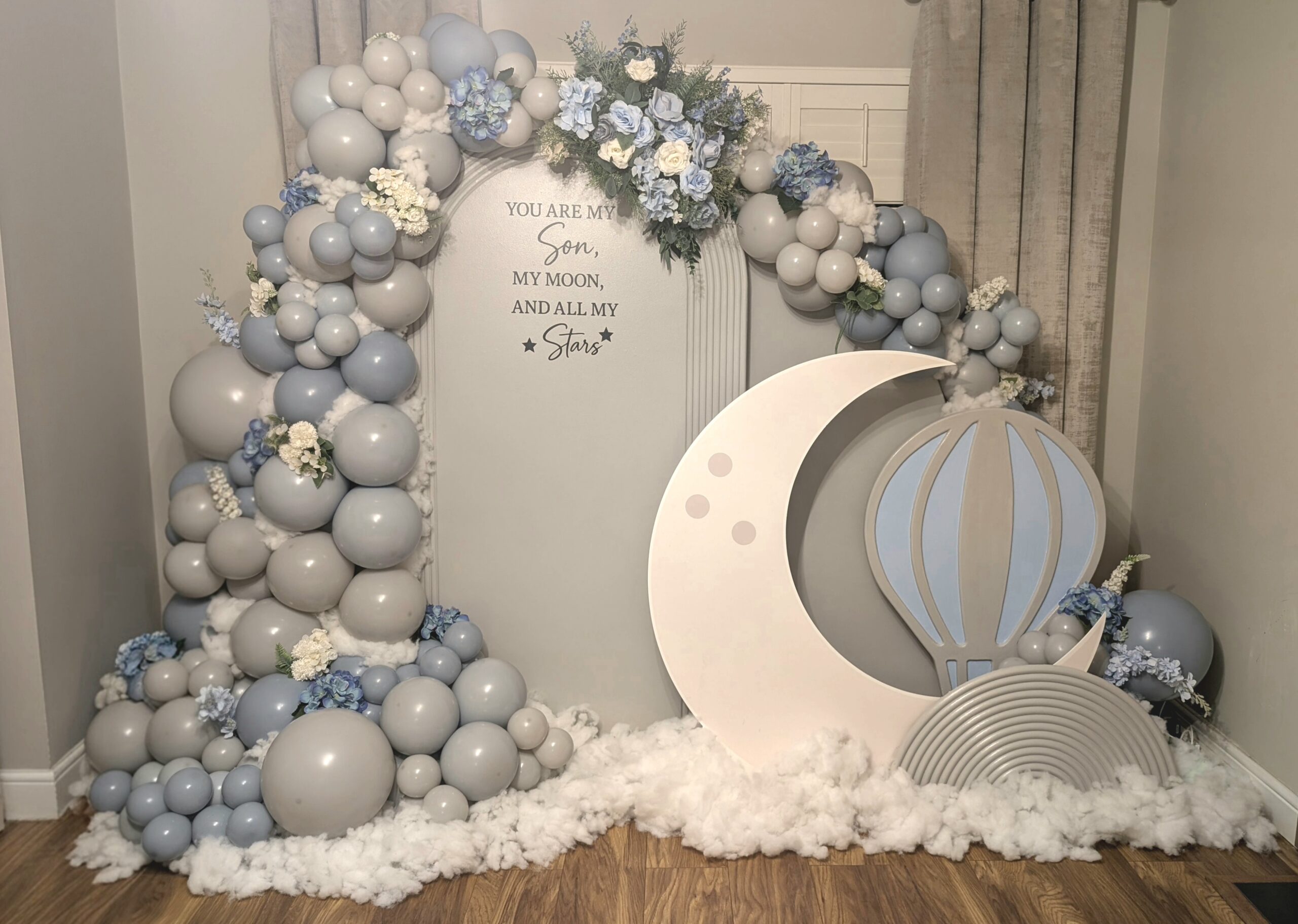 Moon and stars baby backdrop setup with crescent moon and hot air balloon props, balloon arch florals and cloud floor styling