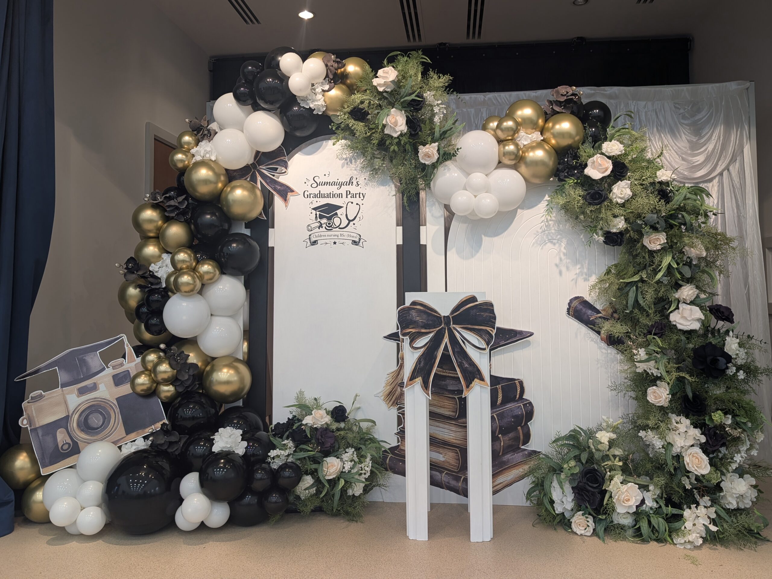 Custom graduation party decor for Sumaiyah with personalised panel, balloon arch in black, white and gold, and greenery accents